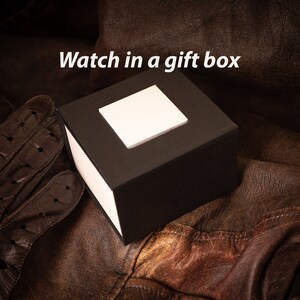 Puede incluir: Una caja de regalo negra con un cuadrado blanco en la parte superior. La caja est&aacute; sobre una superficie de cuero marr&oacute;n.
