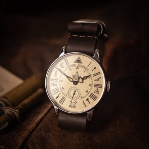 Puede incluir: Un reloj de bolsillo plateado con una correa de cuero marr&oacute;n. La esfera del reloj es de color crema con marcas de n&uacute;meros romanos negros y un dise&ntilde;o de br&uacute;jula en blanco y negro. La esfera del reloj tambi&eacute;n presenta el texto "Мировая Коллекция" en negro.