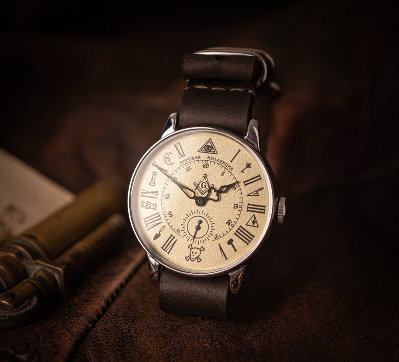 Puede incluir: Un reloj de bolsillo plateado con una correa de cuero marr&oacute;n. La esfera del reloj es de color crema con marcas de n&uacute;meros romanos negros y un dise&ntilde;o de br&uacute;jula en blanco y negro. La esfera del reloj tambi&eacute;n presenta el texto "Мировая Коллекция" en negro.