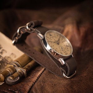 Puede incluir: Un reloj de bolsillo plateado con n&uacute;meros romanos en una correa de cuero marr&oacute;n. La esfera del reloj tiene un fondo crema y presenta un subdial.