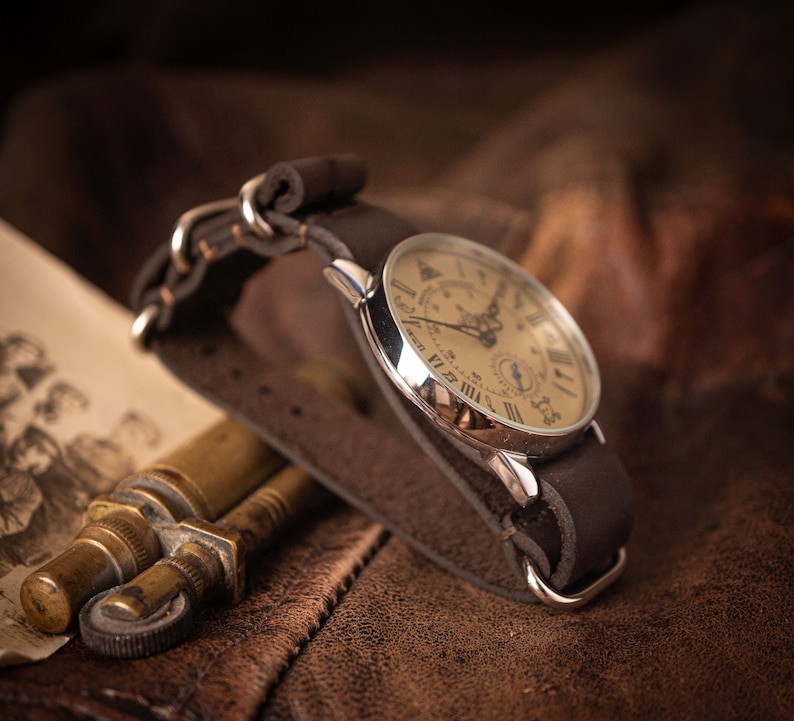 Puede incluir: Un reloj de bolsillo plateado con n&uacute;meros romanos en una correa de cuero marr&oacute;n. La esfera del reloj tiene un fondo crema y presenta un subdial.