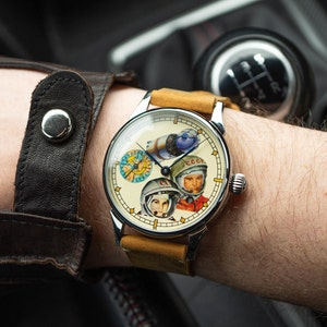 Peut inclure: Une montre-bracelet avec un bracelet en cuir beige et un cadran blanc orné d'une illustration colorée de trois astronautes et du texte "CCCP".