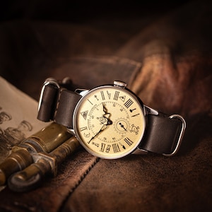 Puede incluir: Un reloj de pulsera de estilo vintage con una esfera de color crema y una correa de cuero negro. La esfera del reloj presenta n&uacute;meros romanos, un peque&ntilde;o subdial y varios s&iacute;mbolos, incluyendo una luna creciente, una estrella y un ojo que todo lo ve.