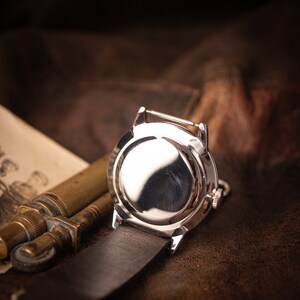 Puede incluir: Un reloj de pulsera plateado con una correa de cuero, visto desde la parte posterior. La esfera del reloj es visible y tiene una superficie reflectante.