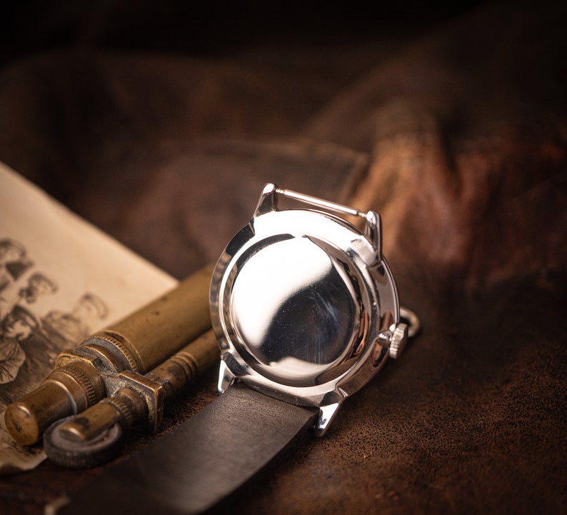Puede incluir: Un reloj de pulsera plateado con una correa de cuero, visto desde la parte posterior. La esfera del reloj es visible y tiene una superficie reflectante.
