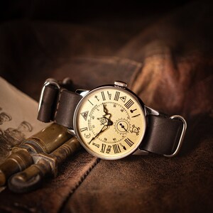 Puede incluir: Un reloj de pulsera de estilo vintage con una esfera de color crema y una correa de cuero negro. La esfera del reloj presenta n&uacute;meros romanos, un peque&ntilde;o subdial y varios s&iacute;mbolos, incluyendo una luna creciente, una estrella y un ojo que todo lo ve.