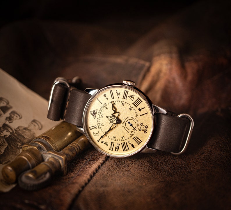 Puede incluir: Un reloj de pulsera de estilo vintage con una esfera de color crema y una correa de cuero negro. La esfera del reloj presenta n&uacute;meros romanos, un peque&ntilde;o subdial y varios s&iacute;mbolos, incluyendo una luna creciente, una estrella y un ojo que todo lo ve.