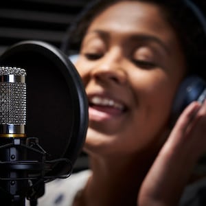 May include: A person singing into a microphone in a recording studio. The microphone is attached to a black pop filter. The person is wearing headphones.