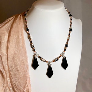 Puede incluir: Un collar de cuentas con tres colgantes negros en forma de diamante. El collar presenta una mezcla de pequeñas cuentas de colores en tonos marrón, transparente y naranja. Los colgantes tienen pequeñas piedras de acento transparentes.
