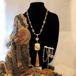 May include: A brown, gray, and beige patterned scarf with a long necklace featuring a large, rectangular, brown, gray, and white stone pendant. The necklace has a tassel and is displayed on a black mannequin. A pair of earrings with a similar stone design are displayed on a stand.