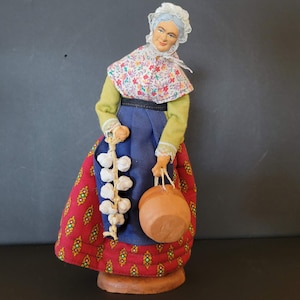 May include: A handcrafted figurine of a woman in traditional attire. She wears a white bonnet, floral shawl, green top, blue skirt, and a red patterned apron. The figurine holds a string of garlic and a brown jug. The base is a light brown color.