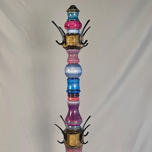 May include: A tall, decorative coat rack with a multi-colored, stacked design. The rack features various shades of blue, pink, purple, and gold, with black metal hooks for hanging items. The wooden structure is topped with a small, dark finial.
