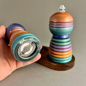May include: A set of two wooden salt and pepper shakers with a multi-colored striped design. The shakers are signed "Tyler Brown 2025" and are sitting on a wooden base.