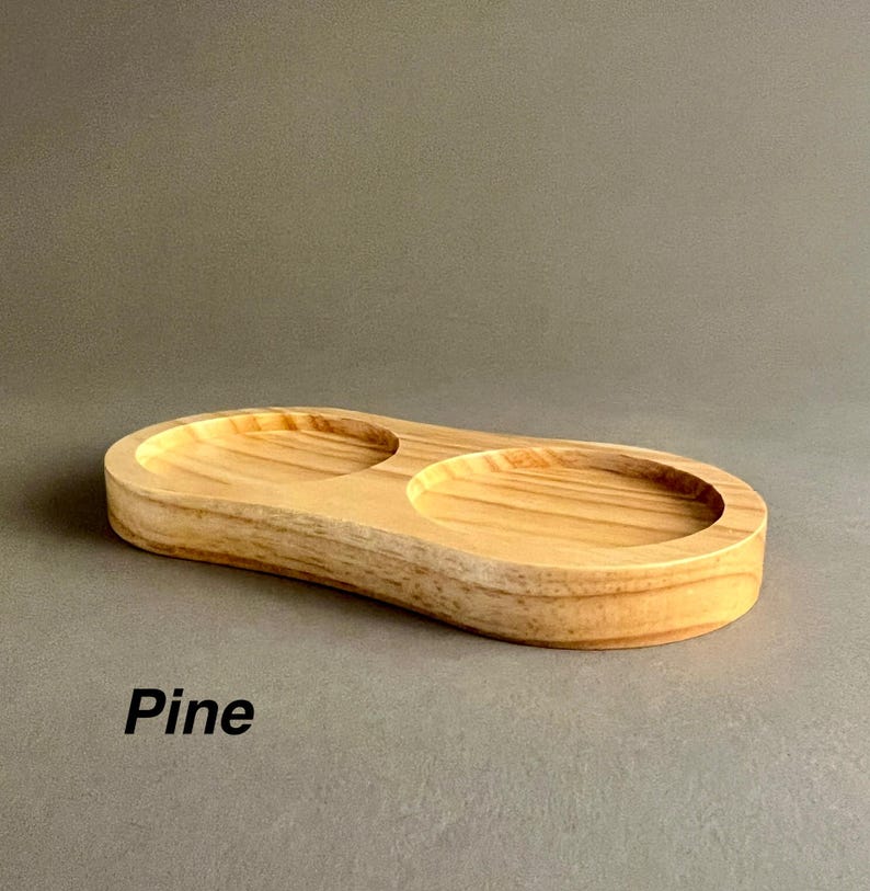 May include: A wooden serving tray with two circular depressions. The tray is made of pine wood and has a natural wood finish. "Pine" is written in white text below the tray.