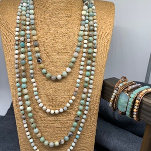 May include: A multi-strand necklace and bracelet set made with green, blue, and white gemstone beads. The necklace is displayed on a brown mannequin bust. The bracelet is wrapped around a wooden stand.