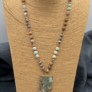 Puede incluir: Un collar largo con cuentas marrones, grises y azules y un gran colgante de piedra verde, marrón y blanca.