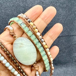 Op de afbeelding: Een meerstrengs wikkelarmband met een grote, ovale, lichtgroene stenen hanger. De armband bevat verschillende kralen in groen-, bruin- en wittinten, met metallic accenten. De armband is om de pols gewikkeld.