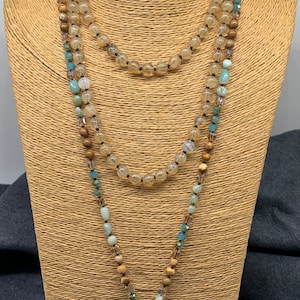 May include: A three-strand necklace displayed on a mannequin. The necklace features clear and brown beads, with accents of blue and brown stones. The pendant is a large, teardrop-shaped turquoise stone set in a gold-colored frame.