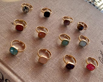 May include: A collection of 12 gold rings with square-shaped gemstones in various colors, including turquoise, pink, red, black, and green.