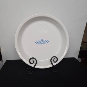 May include: White ceramic pie plate with a blue floral design in the center. The plate is displayed on a black metal stand with decorative scrollwork. The plate has a slightly raised rim.