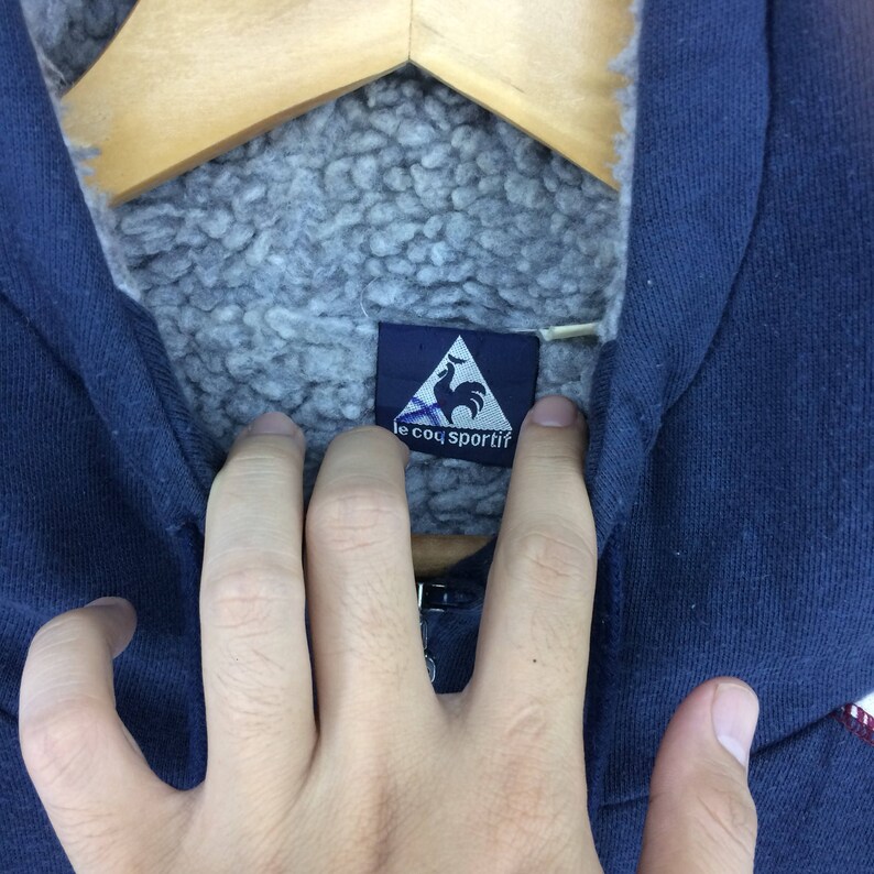 K&ouml;nnte beinhalten: Nahaufnahme eines marineblauen Kapuzenpullovers mit grauem, flauschigem Innenfutter. Das Etikett zeigt ein wei&szlig;es Dreieck mit einem Hahn-Logo und dem Text "le coq sportif".