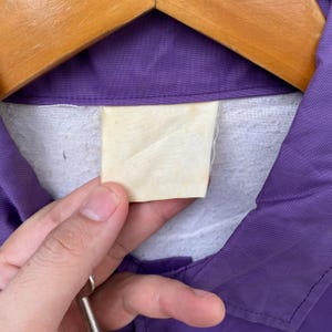 May include: A purple work shirt with a white label attached to the collar. The label is blank.