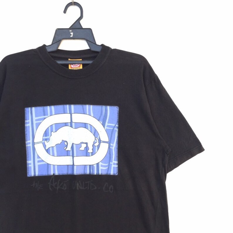 May include: Black t-shirt with a white rhino graphic inside a blue and white plaid design. The shirt has the text "THE Ecko UNLTD. CO." printed below the graphic. The shirt is hanging on a black hanger.