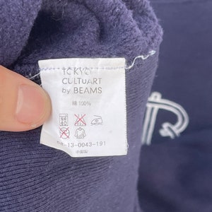 May include: A white tag with black text that reads "Tokyo Cultuart by Beams" and "綿 100%". The tag also includes care instructions and the number "58-13-0043-191". The tag is attached to a dark blue garment.