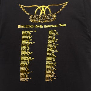 May include: A black t-shirt with a gold Aerosmith logo and the text "Nine Lives North American Tour". The shirt lists cities and states where the band will be performing on their tour.