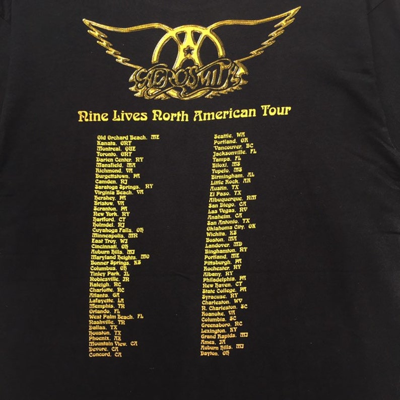 May include: A black t-shirt with a gold Aerosmith logo and the text "Nine Lives North American Tour". The shirt lists cities and states where the band will be performing on their tour.