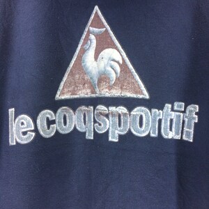 K&ouml;nnte beinhalten: Marineblaues Sweatshirt mit dem Le Coq Sportif-Logo. Das Logo zeigt einen wei&szlig;en Hahn in einem braunen Dreieck. Der Markenname "le coq sportif" ist in silberner, strukturierter Schrift darunter gedruckt.