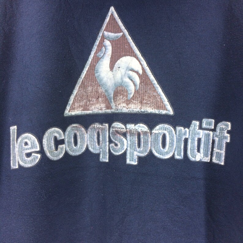 K&ouml;nnte beinhalten: Marineblaues Sweatshirt mit dem Le Coq Sportif-Logo. Das Logo zeigt einen wei&szlig;en Hahn in einem braunen Dreieck. Der Markenname "le coq sportif" ist in silberner, strukturierter Schrift darunter gedruckt.