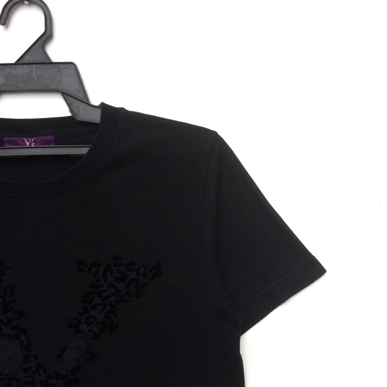 May include: Black crew neck t-shirt with a dark floral design. The shirt is hanging on a black hanger. The brand name "Y's" is visible on a purple tag.