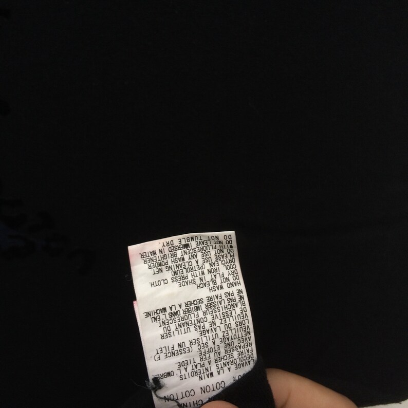 May include: A black garment with a white tag attached. The tag has text in multiple languages, including French and English, detailing care instructions such as hand wash, dry flat, and cool iron.