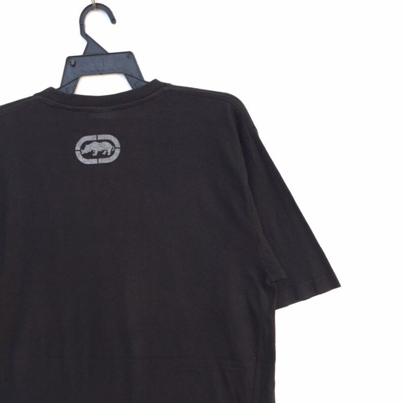 May include: Black short-sleeved t-shirt with a gray logo on the back. The logo is a stylized rhinoceros inside an oval. The shirt is hanging on a black hanger against a white background.