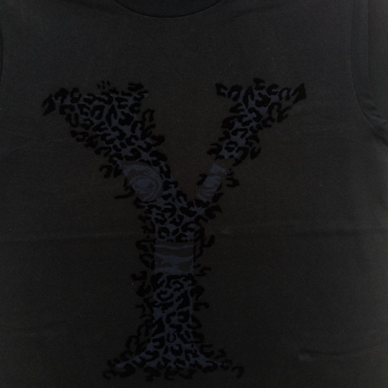 May include: Black t-shirt featuring a large, stylized letter "Y" in a dark blue leopard print design. The letter incorporates floral elements and a band detail, creating a unique graphic.