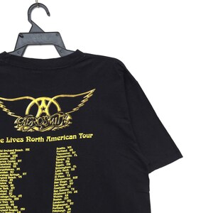 May include: Black t-shirt with a gold Aerosmith logo and the text "e Lives North American Tour" with a list of cities and states where the band will be performing.
