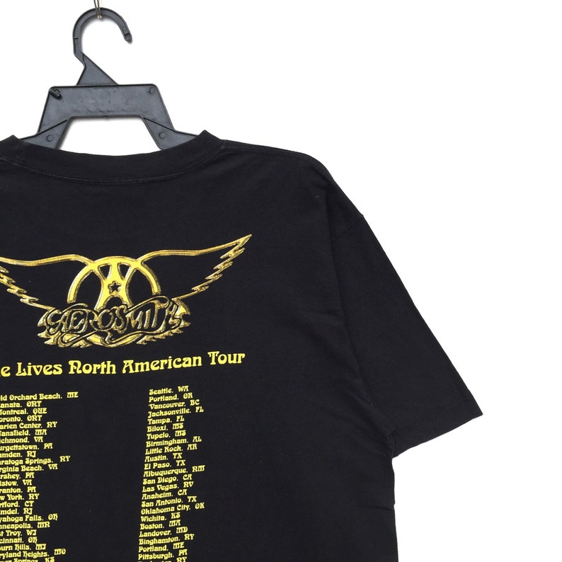 May include: Black t-shirt with a gold Aerosmith logo and the text "e Lives North American Tour" with a list of cities and states where the band will be performing.