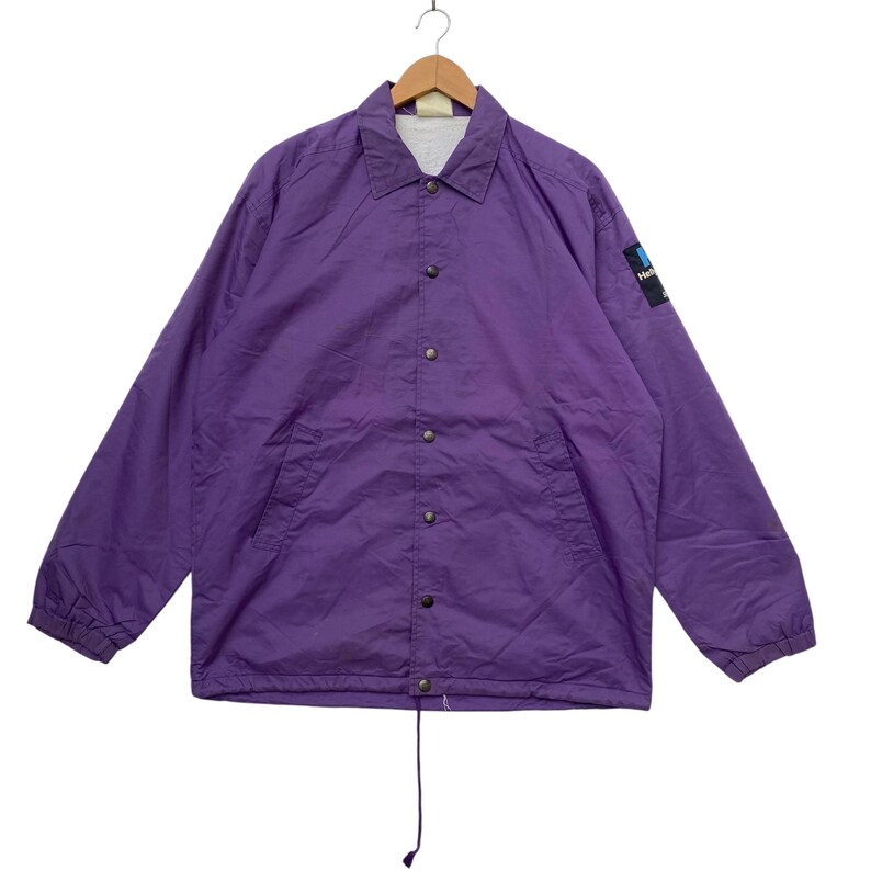 May include: A purple nylon coach jacket with a snap closure and a small logo on the left sleeve. The jacket has a drawstring at the bottom.