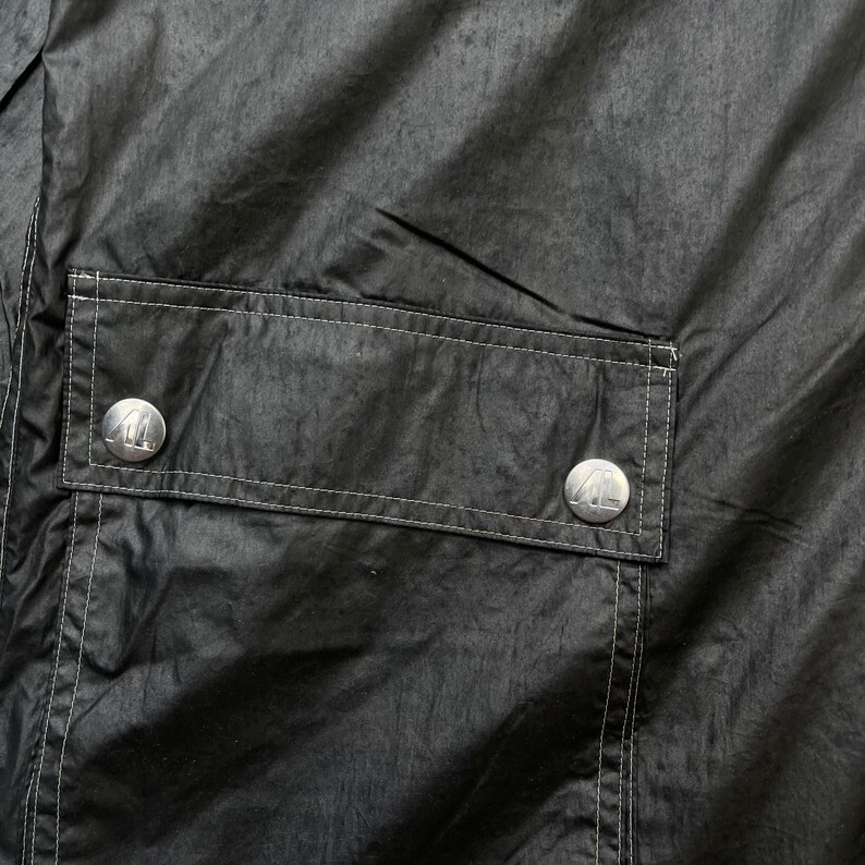 May include: A black jacket with a large pocket on the front. The pocket has two silver buttons with a logo on them.