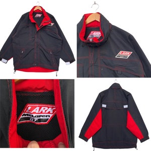 Może przedstawiać: Czarna i czerwona kurtka z logo Lark McLaren GT. Kurtka ma zamek błyskawiczny, dwie kieszenie i kaptur ze sznurkiem.