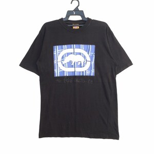 May include: Black t-shirt featuring a rectangular graphic with a white rhino inside a white oval, set against a blue plaid background. The text "the Ecko UNLTD. CO" is below the graphic. The shirt is hanging on a black hanger.