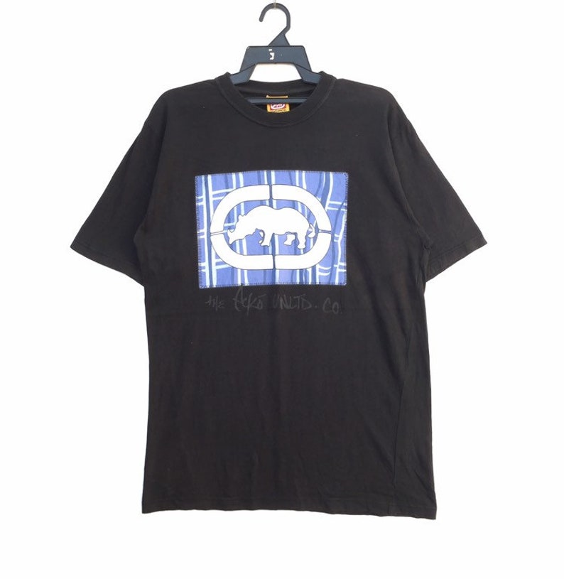 May include: Black t-shirt featuring a rectangular graphic with a white rhino inside a white oval, set against a blue plaid background. The text "the Ecko UNLTD. CO" is below the graphic. The shirt is hanging on a black hanger.
