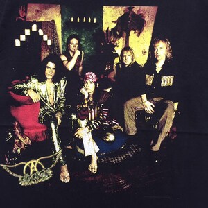 May include: A vintage band t-shirt featuring a black and white photo of five people in a dimly lit room. The band's name, "Aerosmith", is printed in gold with a winged logo.