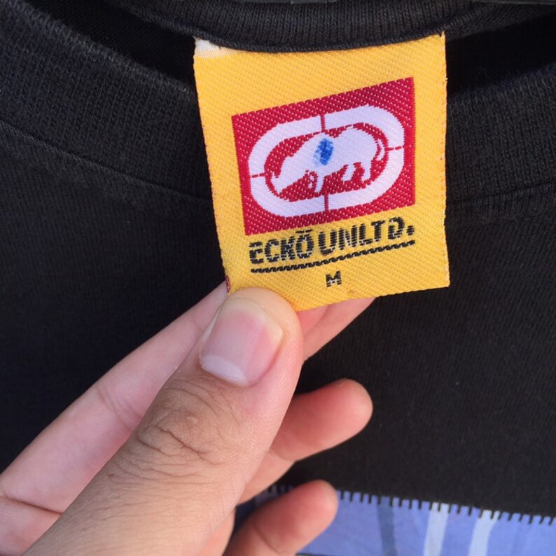 May include: Close-up of a yellow and red Ecko Unltd. tag on a black shirt. The tag features the Ecko Unltd. logo, a white rhinoceros inside a red target, and the text "ECKO UNLTD." and "M".