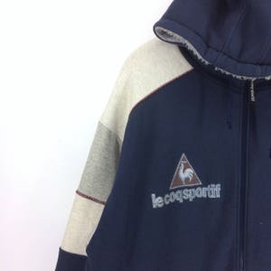 K&ouml;nnte beinhalten: Marineblaue Kapuzenjacke mit Rei&szlig;verschluss und einem Colorblock-Design in Grau und Off-White. Mit dem gestickten Logo eines Hahns in einem Dreieck und dem Text "le coq sportif". Die Kapuze hat ein weiches, helles Futter.