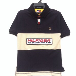 Può includere: Una polo a righe blu navy e crema con una patch logo rosso e bianco che dice "Hilfiger Alpine Gear" sul davanti. La polo ha un piccolo logo ricamato sul petto destro.