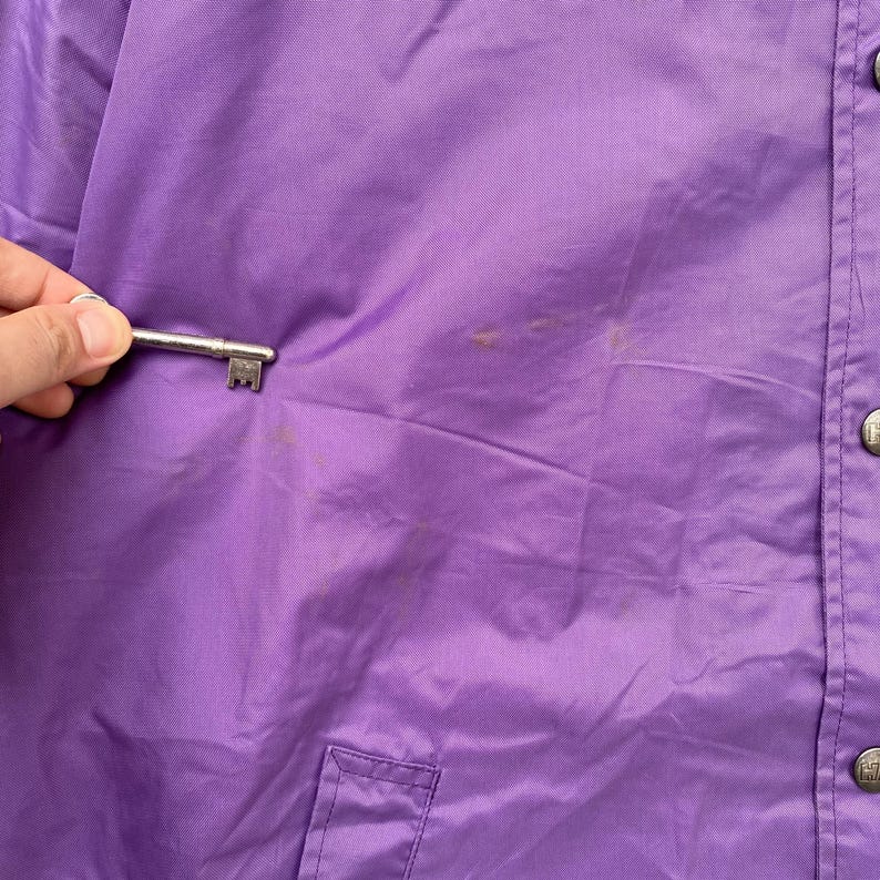 May include: A purple nylon jacket with a button closure and a pocket on the left side. The jacket has a few light stains.