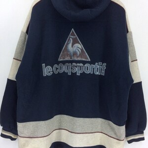 K&ouml;nnte beinhalten: Ein marineblauer Kapuzenpullover mit einem grau-cremefarbenen Farbblock-Design. Auf der R&uuml;ckseite befindet sich ein Dreieckslogo mit einem Hahn und dem Text "le coq sportif". Der Pullover hat ein Fleecefutter und eine Kapuze mit Kordelzug.