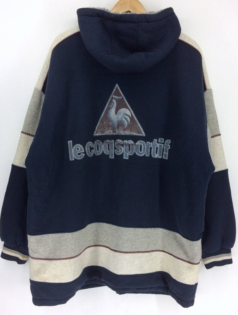 K&ouml;nnte beinhalten: Ein marineblauer Kapuzenpullover mit einem grau-cremefarbenen Farbblock-Design. Auf der R&uuml;ckseite befindet sich ein Dreieckslogo mit einem Hahn und dem Text "le coq sportif". Der Pullover hat ein Fleecefutter und eine Kapuze mit Kordelzug.